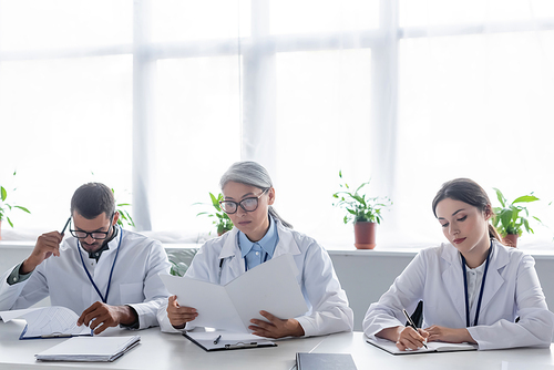 middle aged asian physician looking in paper folder near young colleagues during meeting