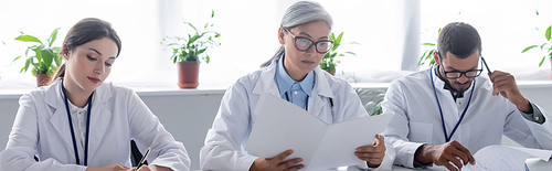 thoughtful doctor touching head with pen while working near multiethnic colleagues, banner