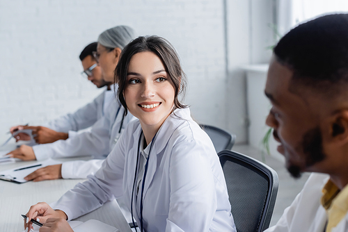 young doctor smiling at african american colleagues near physicians on blurred background