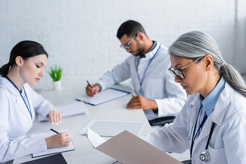 mature asian doctor looking in paper folder near blurred colleagues working with papers