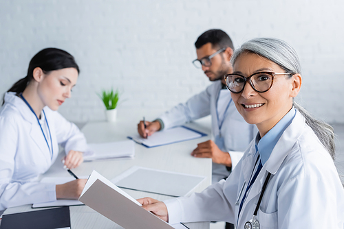 middle aged asian doctor in eyeglasses smiling at camera near young colleagues on blurred background