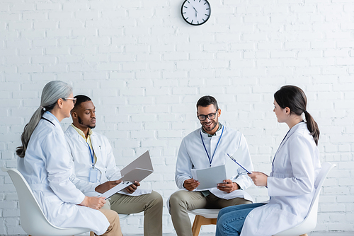 young doctor with papers smiling near multiethnic colleagues during medical council