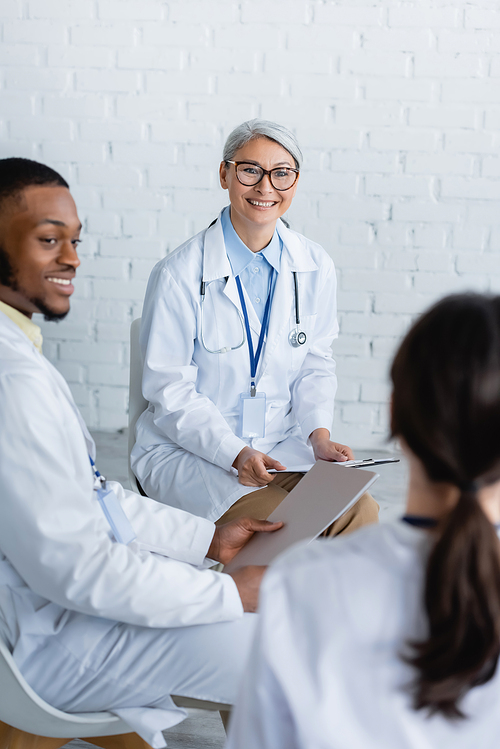 mature asian doctor in eyeglasses smiling near blurred multiethnic colleagues during council