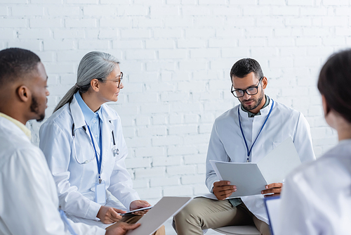 doctor in eyeglasses looking at documents near multiethnic colleagues