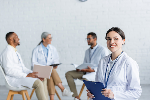 young doctor with clipboard smiling at camera while multiethnic colleagues talking on blurred background