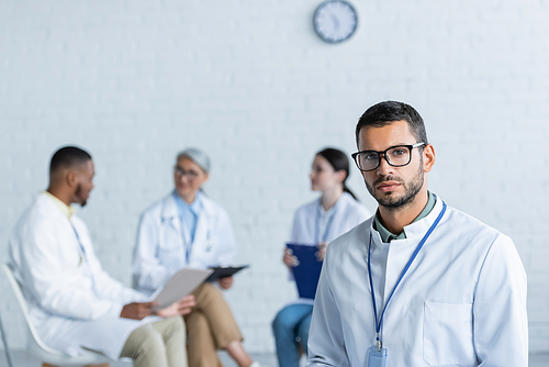 young physician in eyeglasses  while multiethnic doctors talking on blurred background