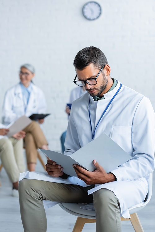 young doctor in eyeglasses looking in paper folder near blurred colleagues