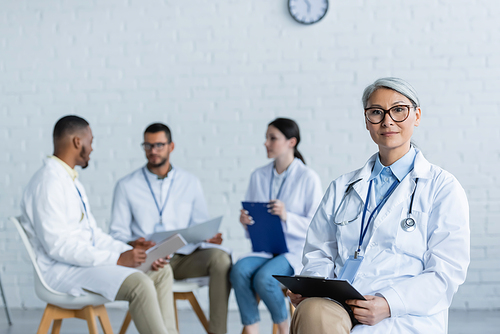 middle aged asian physician with clipboard  near multiethnic colleagues talking on blurred background