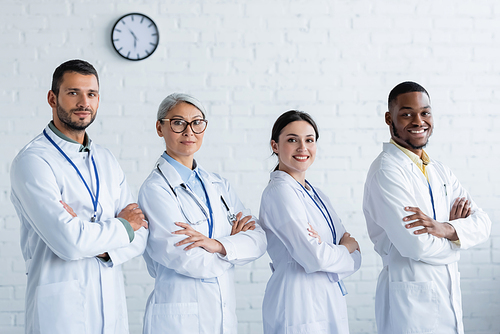 smiling multiethnic doctors in white coats smiling at camera while standing with crossed arms