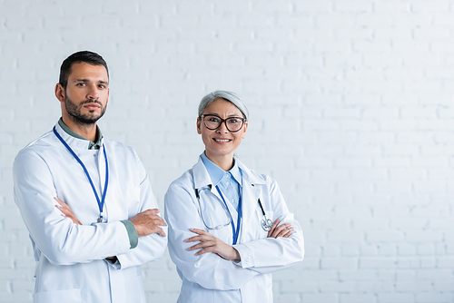 asian doctor smiling near serious colleague while standing with crossed arms