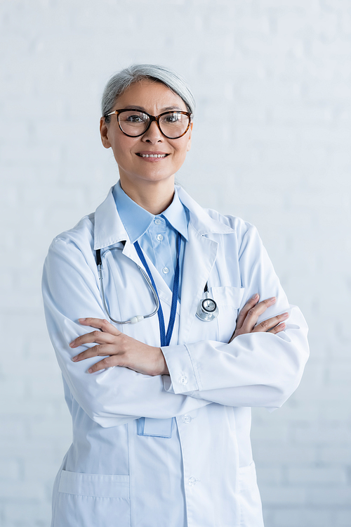 mature asian doctor in white coat and eyeglasses standing with crossed arms and smiling at camera on white