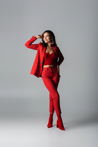full length of young happy woman in red boots and suit posing on grey