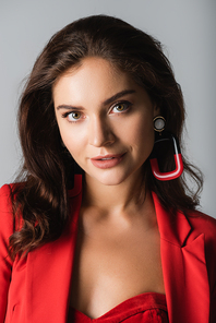 pleased young trendy woman in red suit and earrings posing isolated on grey