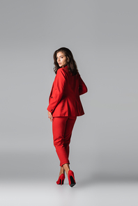 full length of young stylish woman in red heels and suit posing while walking on grey