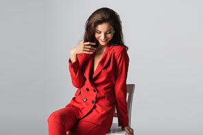 cheerful young woman in red suit posing while sitting on white chair isolated on grey