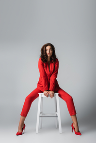 full length of brunette woman in red suit posing while sitting on white chair on grey