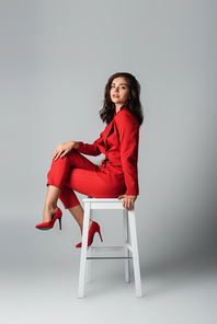 full length of trendy young woman in red suit sitting on white chair on grey