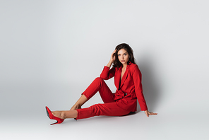 full length of trendy woman in red suit posing while sitting on grey