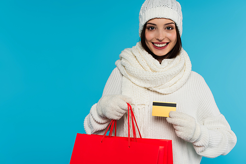 Pretty woman in hat and gloves holding shopping bags and credit card isolated on blue