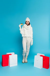 Smiling woman in warm clothes and gloves holding credit card near shopping bags on blue background
