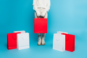 Cropped view of woman in sweater and gloves holding shopping bag near purchases on blue background