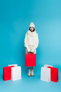 Pretty shopaholic in knitted clothes standing near shopping bags on blue background