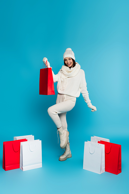 Positive woman in warm hat and gloves holding shopping bags on blue background