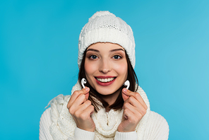 Pretty woman in knitted hat and scarf holding wireless earphones isolated on blue