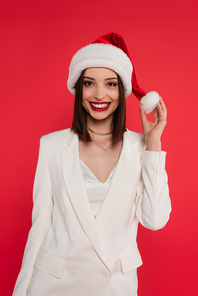 Positive woman in white jacket holding santa hat isolated on red