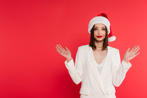 Stylish woman with red lips and santa hat pointing with hands isolated on red