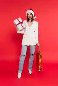 Cheerful woman in santa hat holding shopping bag and present on red background