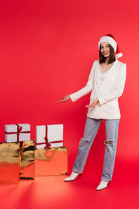 Smiling woman in santa hat pointing at gifts in shopping bags on red background