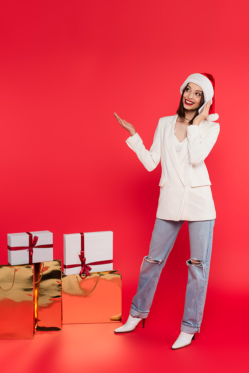 Smiling woman in santa hat talking on smartphone near gifts and shopping bags on red background