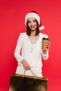 Stylish woman in santa hat holding shopping bag and coffee to go isolated on red