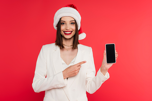 Cheerful woman in santa hat pointing at smartphone with blank screen isolated on red