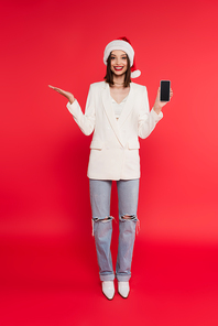Cheerful woman in santa hat and white jacket holding smartphone with blank screen and pointing with hand on red background