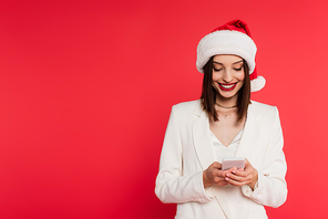 Positive woman in santa hat and white jacket using mobile phone isolated on red