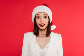 Amazed woman in jacket and santa hat  isolated on red