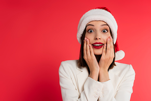Astonished woman with red lips and santa hat looking at camera isolated on red