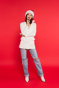Full length of pensive woman in santa hat and white jacket on red background
