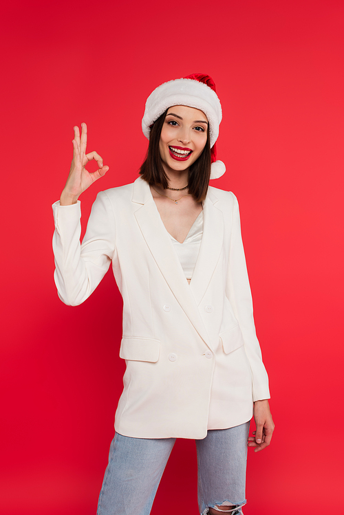 Stylish woman in santa hat showing okay gesture isolated on red
