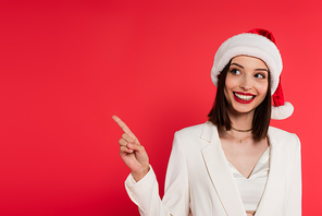 Cheerful woman in santa hat pointing with finger isolated on red