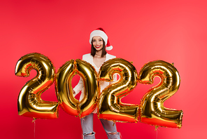 Pretty woman in santa hat standing near balloons in shape of 2022 numbers isolated on red