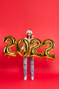 Young woman in santa hat standing near balloons in shape of 2022 numbers on red background