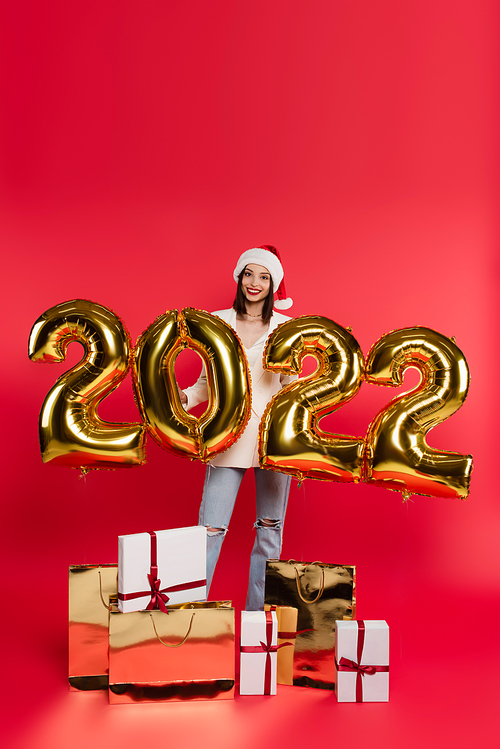 Happy woman in santa hat standing near balloons in shape of 2022 numbers, gifts and shopping bags on red background