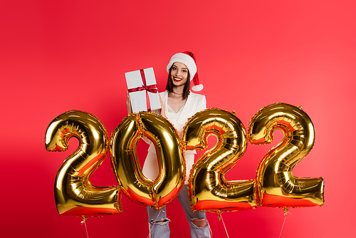 Smiling woman in santa hat holding present near balloons in shape of 2022 isolated on red