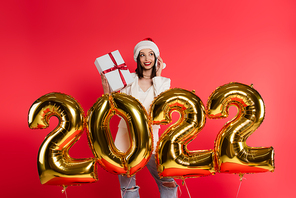 Smiling woman in santa hat talking on smartphone and holding gift near balloons in shape of 2022 numbers isolated on red