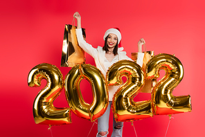 Cheerful woman in santa hat holding shopping bags near balloons in shape of 2022 numbers isolated on red