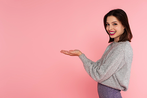 Happy woman in warm sweater pointing with hands isolated on pink