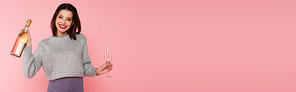 Positive woman holding bottle of champagne and glass isolated on pink, banner